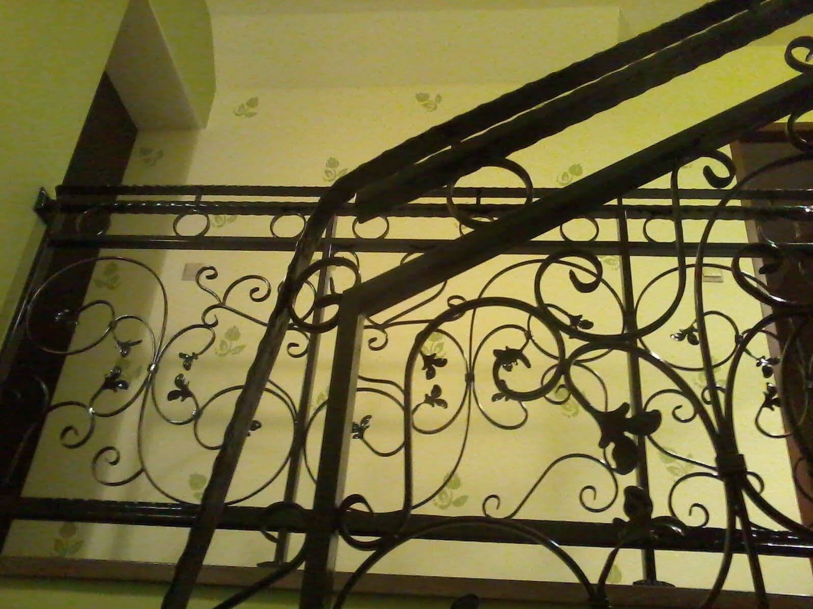 Kuta, czarna balustrada schodowa z motywami roślinnymi na tle ściany w pastelowym odcieniu z delikatnym wzorem. Ujęcie z dołu, widoczne fragmenty schodów.