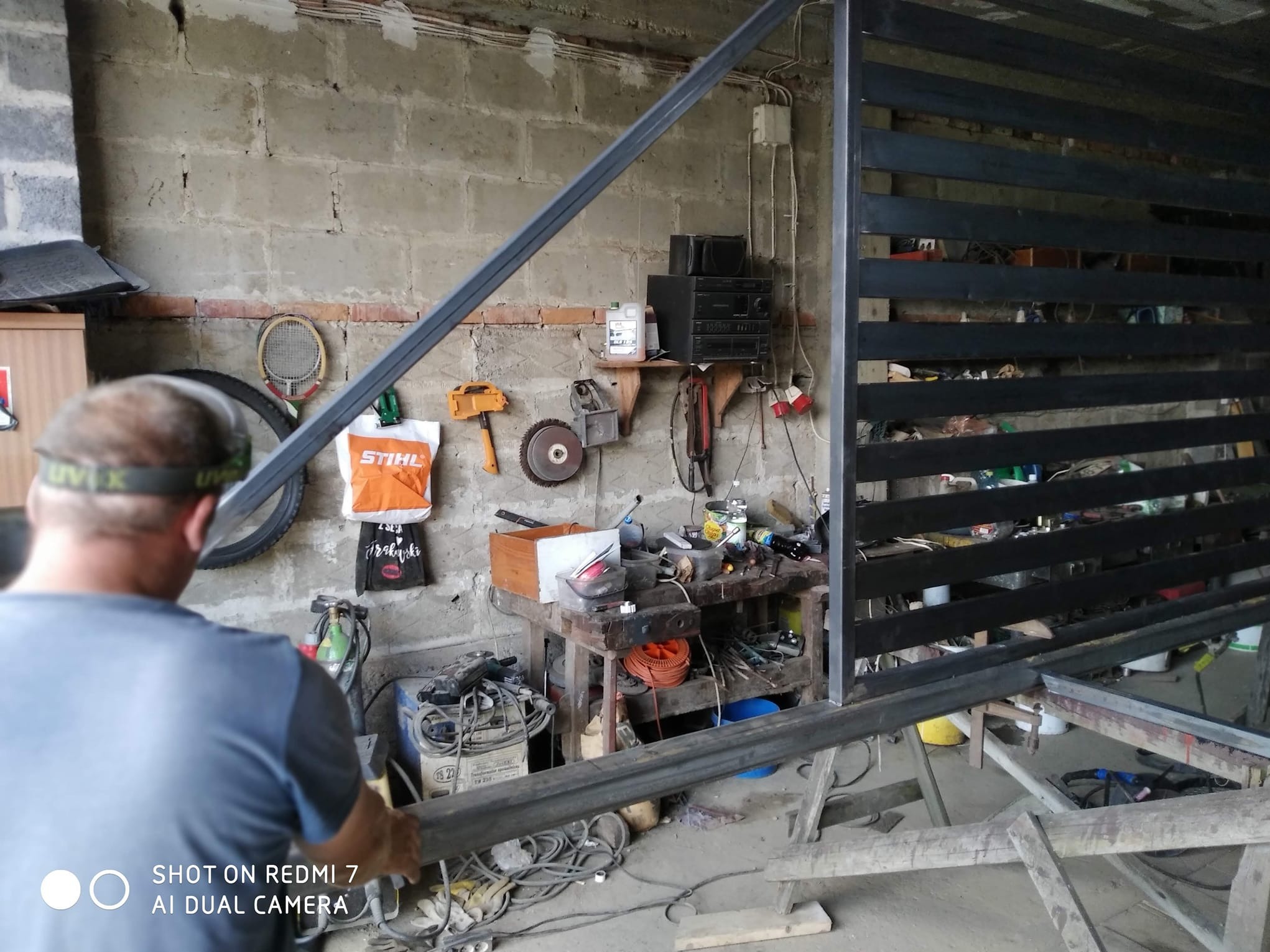 Spawanie metalowej konstrukcji ogrodzenia w warsztacie. Widoczny spawacz przy pracy, narzędzia i elementy ogrodzenia w surowym stanie.