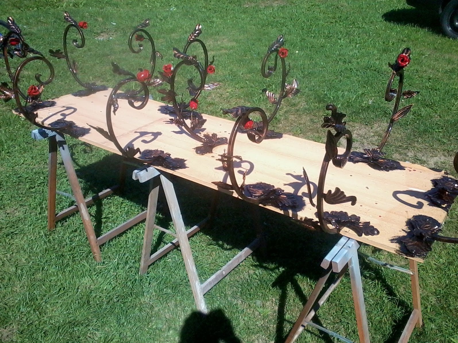 Kute, metalowe elementy dekoracyjne w kształcie roślin z czerwonymi akcentami, ułożone na drewnianej płycie na tle zielonej trawy. Elementy są w trakcie obróbki lub prezentacji.