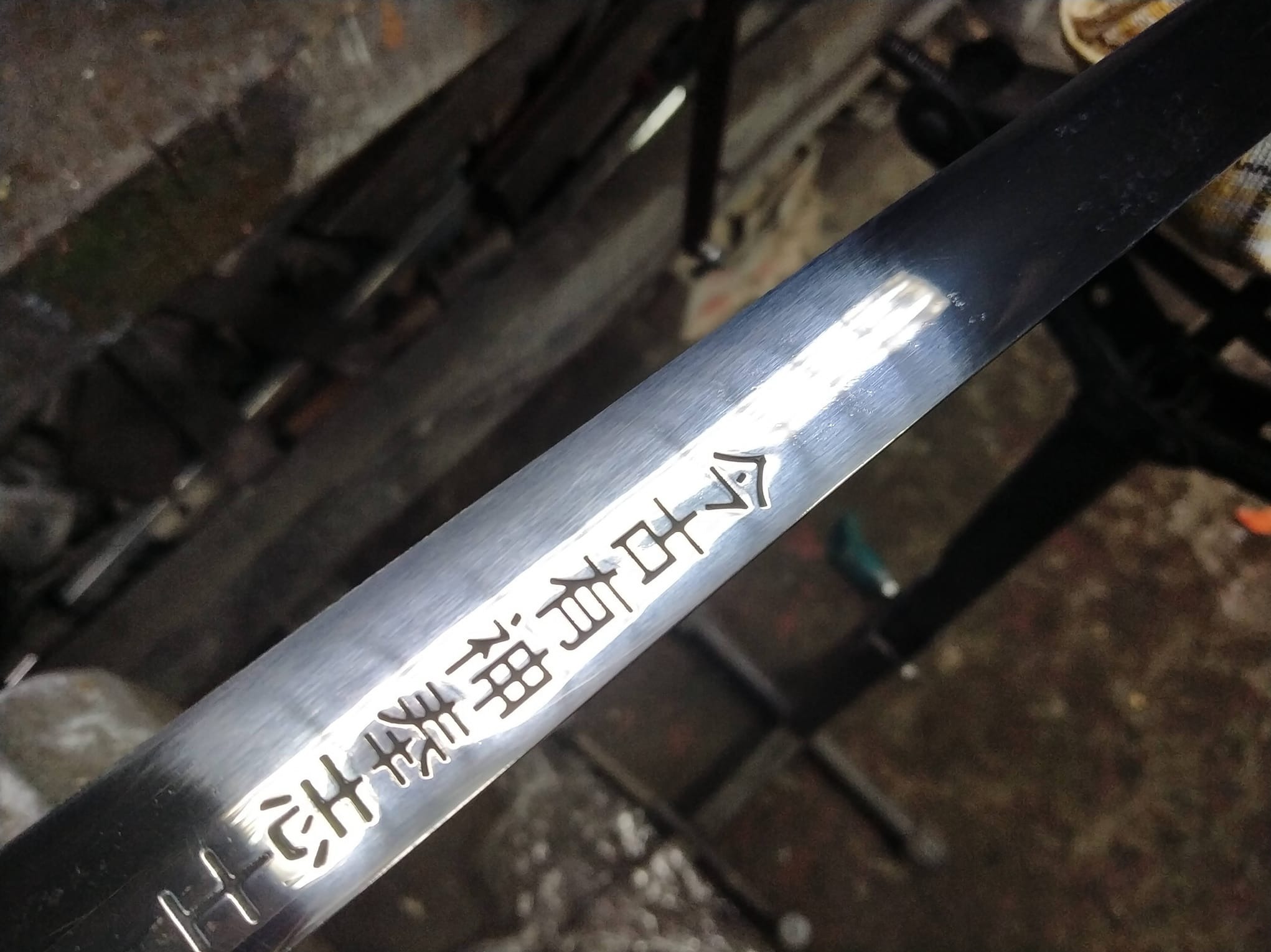 Błyszczące, metalowe ostrze z wygrawerowanymi chińskimi znakami, leżące na warsztacie ślusarskim, uchwycone pod kątem.