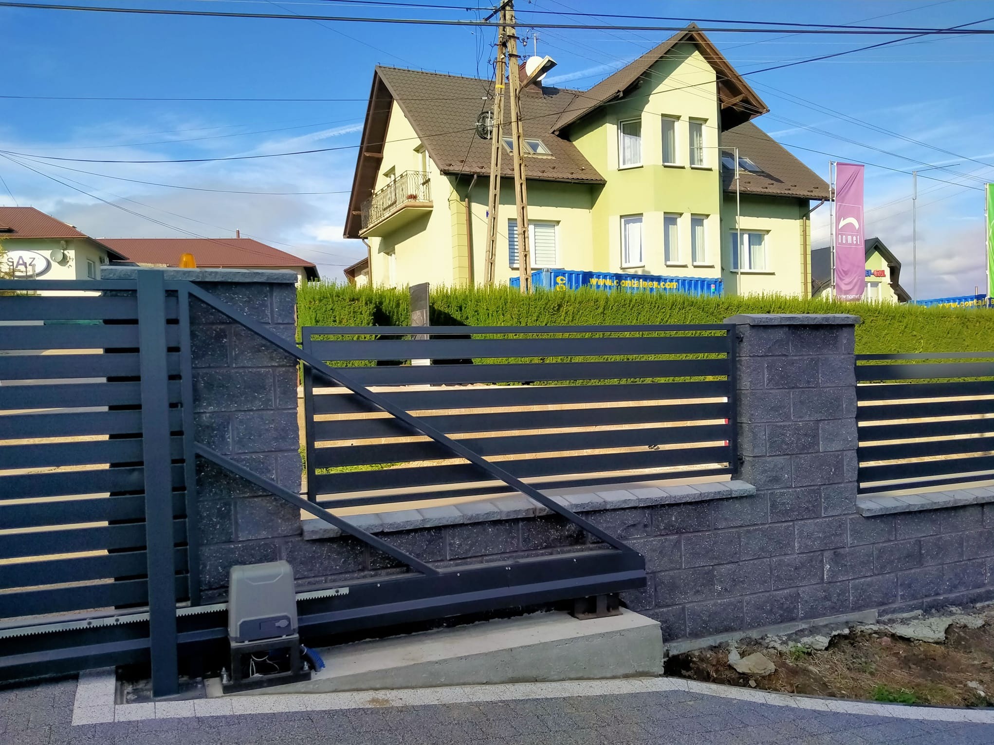 Nowoczesne ogrodzenie metalowe z automatyczną bramą przesuwną, szare słupki z bloczków betonowych, w tle dom z zielonym żywopłotem i błękitne niebo.