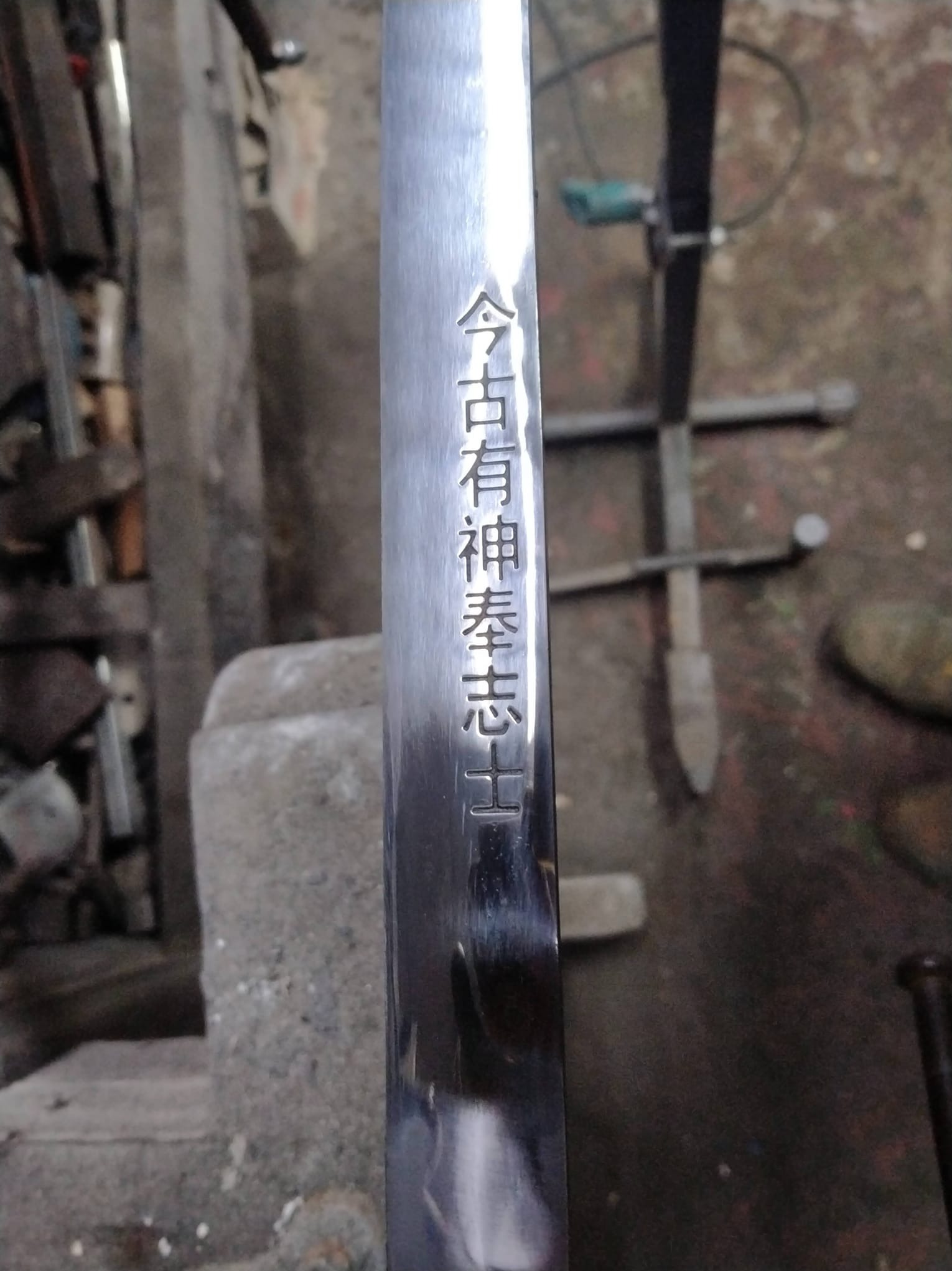 Metalowy element z wygrawerowanymi znakami chińskimi, leżący na warsztacie ślusarskim. Widoczne narzędzia i elementy konstrukcyjne w tle.