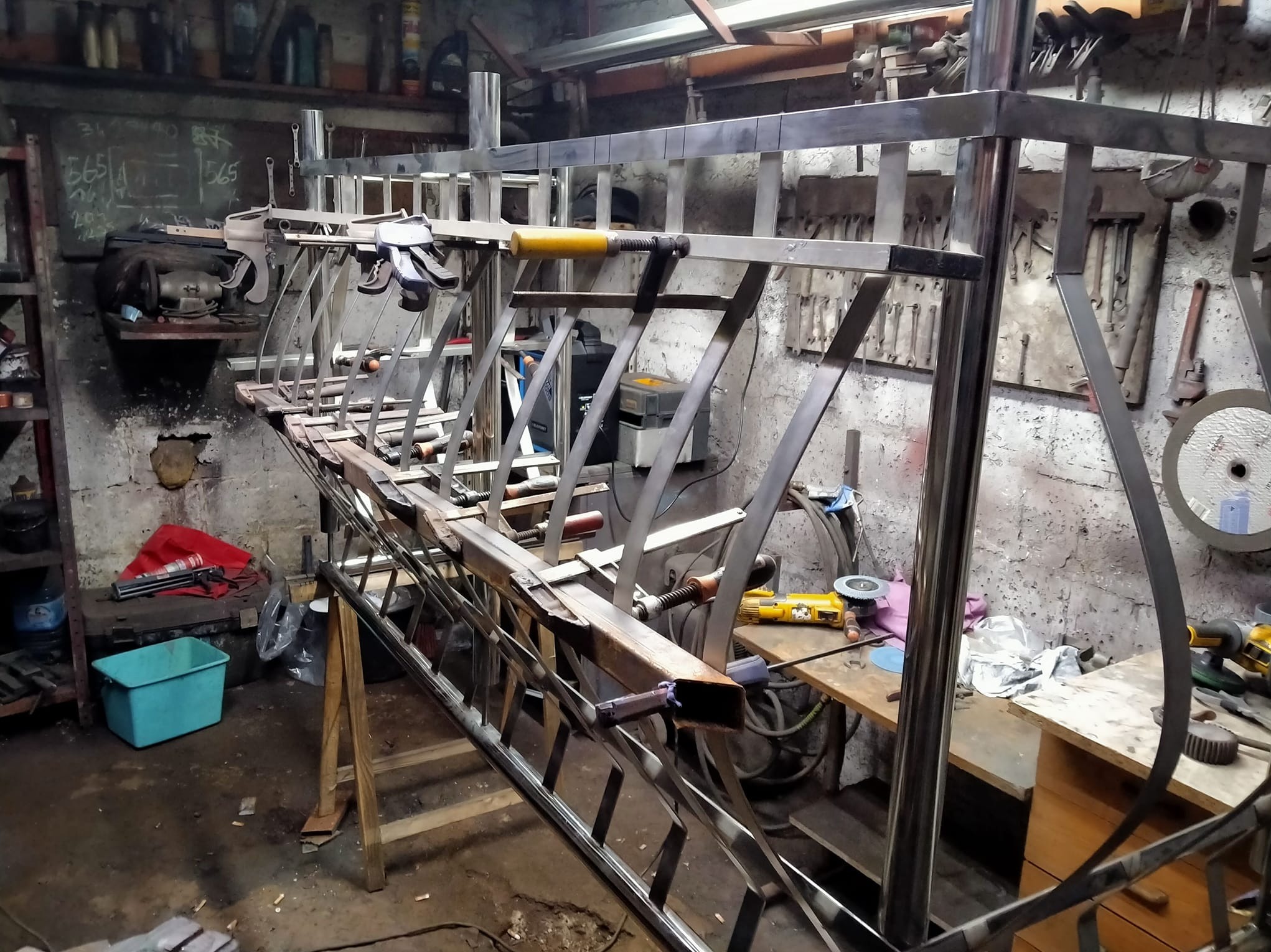 Konstrukcja metalowa w warsztacie, prawdopodobnie balustrada, w trakcie spawania. Widoczne narzędzia i akcesoria ślusarskie. Surowe wykończenie, warsztatowy nieład.