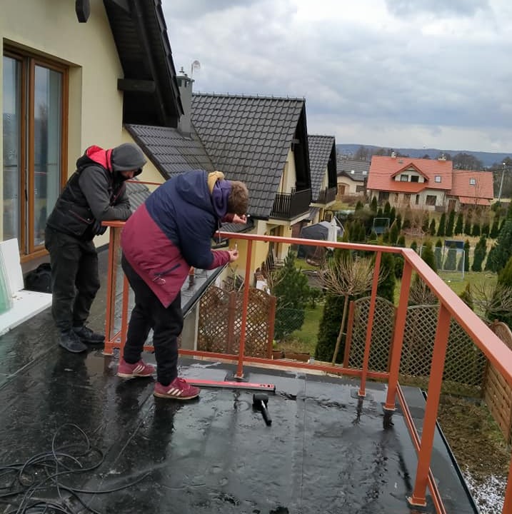 Montaż metalowej balustrady na balkonie z czarną hydroizolacją. Dwóch monterów w trakcie pracy, w tle zabudowa jednorodzinna i pochmurne niebo.