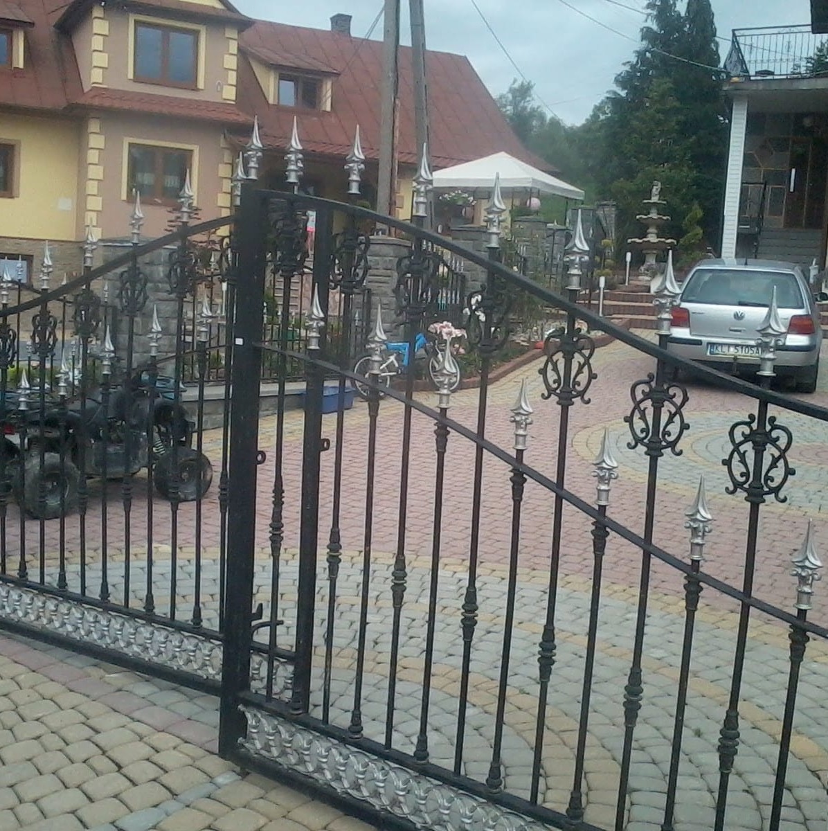 Otwarta, czarna, metalowa brama z dekoracyjnymi elementami w kształcie grotów i rozet na tle posesji z domem, quadem i samochodem osobowym.