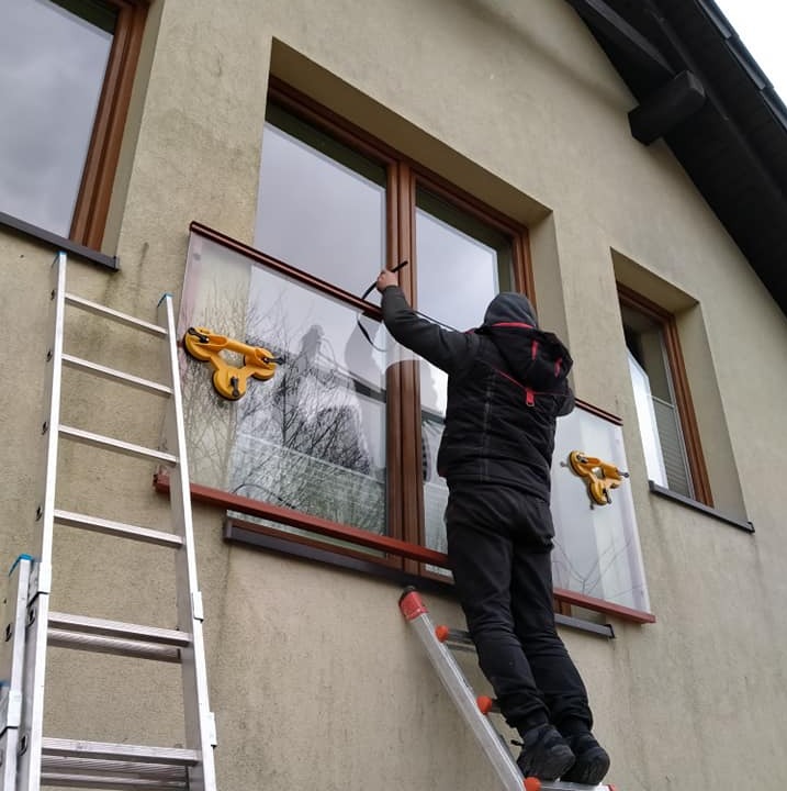 Montaż okna z użyciem przyssawek, widoczna drabina oparta o elewację budynku. Osoba w kurtce montuje szybę w brązowej ramie okiennej.