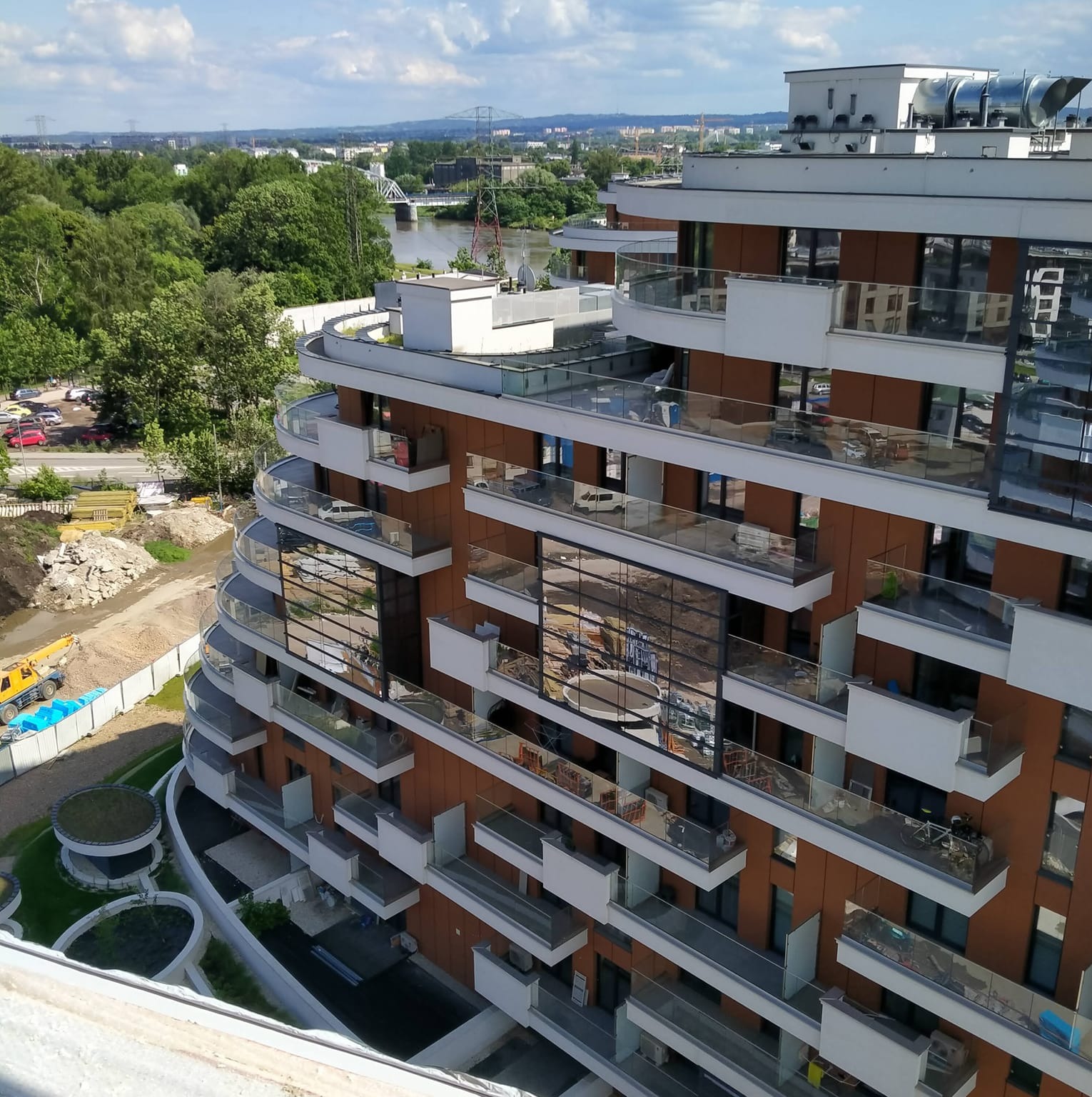 Nowoczesny budynek z balkonami o szklanych balustradach, widok z góry na tle zieleni i rzeki. Architektura z elementami brązu i bieli w słoneczny dzień.