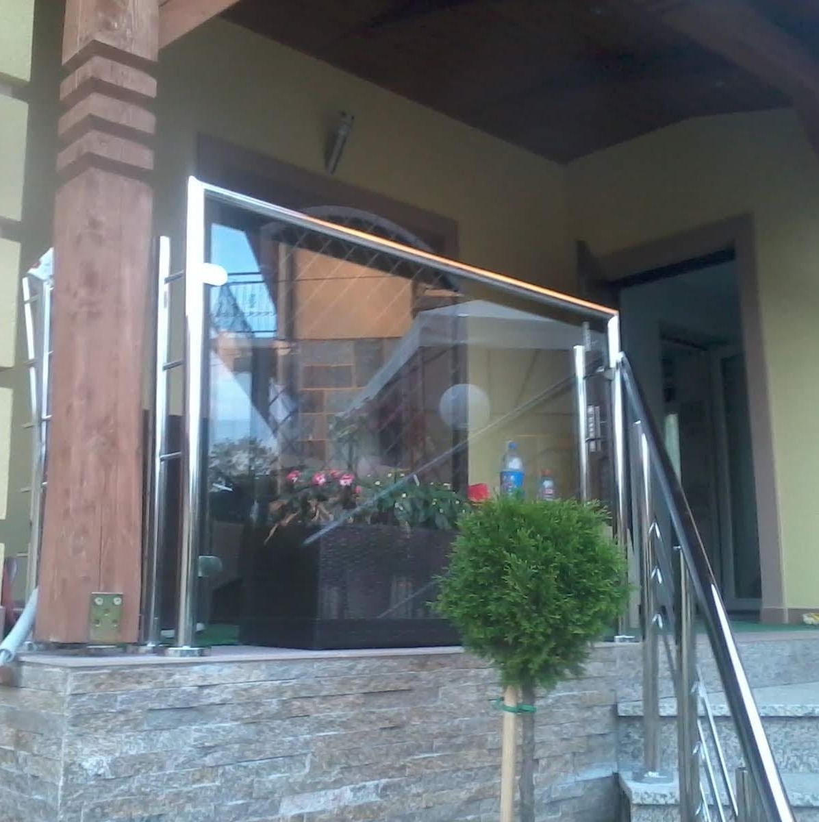 Szklana balustrada ze stali nierdzewnej na tarasie domu. Widoczne schody z metalową poręczą i dekoracyjna roślina. Ujęcie z dołu, podkreślające nowoczesny design.