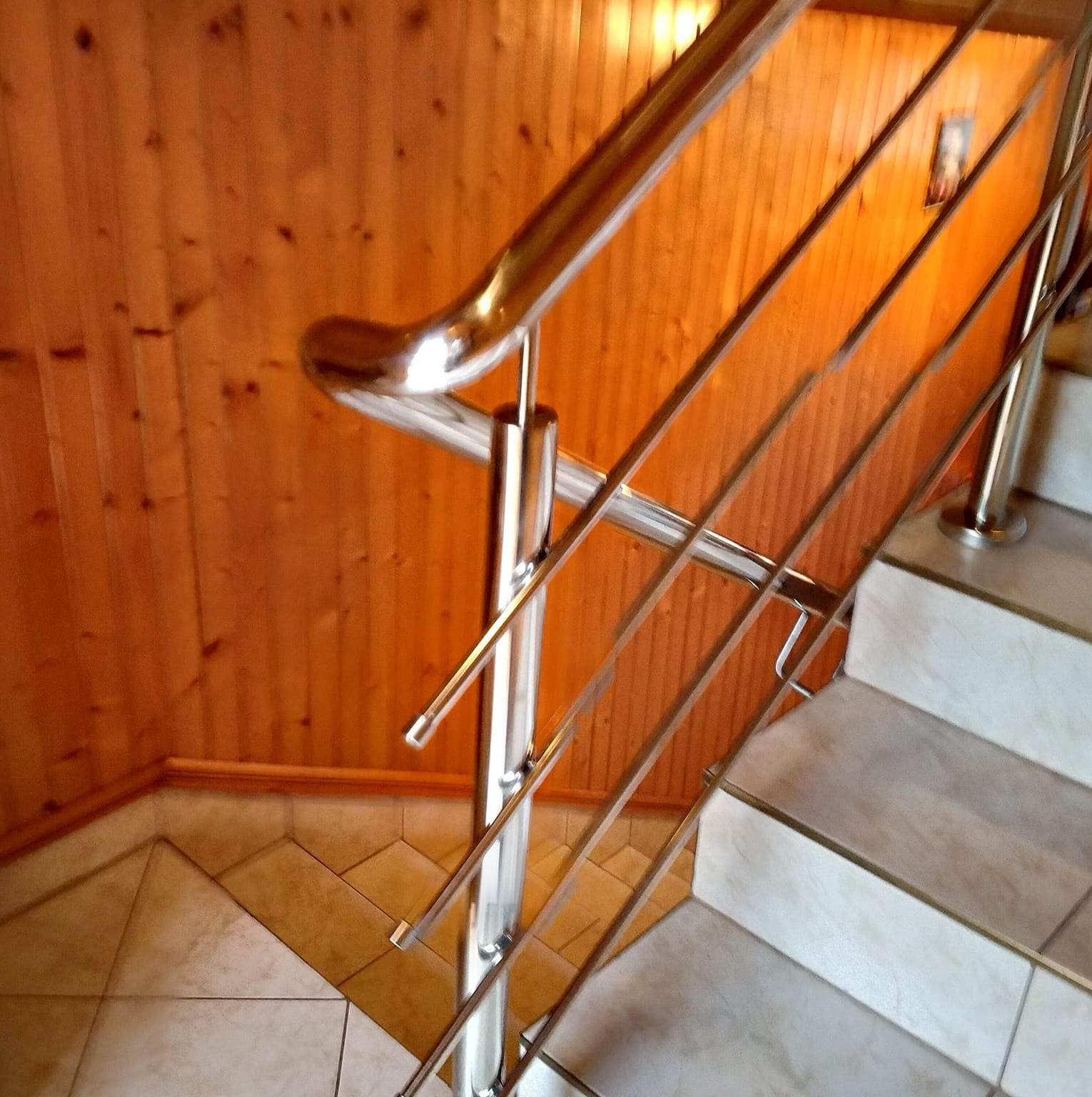 Polerowana balustrada nierdzewna przy schodach z płytek, drewniana boazeria na ścianie w tle. Ujęcie z góry, lekko z ukosa.