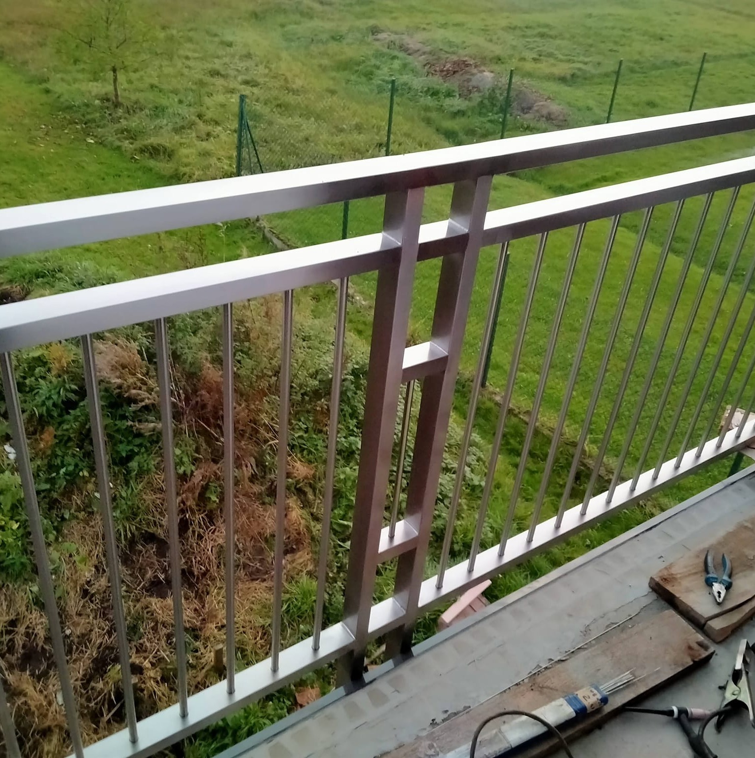 Nowoczesna balustrada balkonowa ze stali nierdzewnej z widokiem na zielony ogród. Widoczne narzędzia i materiały spawalnicze.