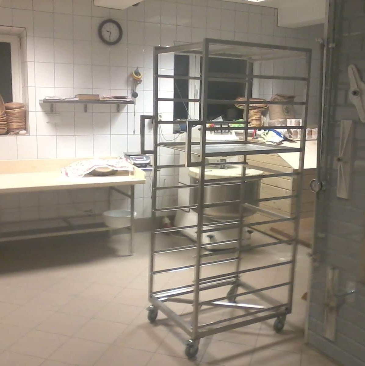 Wnętrze piekarni z metalowym regałem na kółkach, blatem roboczym i piecem. Białe płytki na ścianach, widoczne akcesoria piekarskie. Nierównomierne oświetlenie.