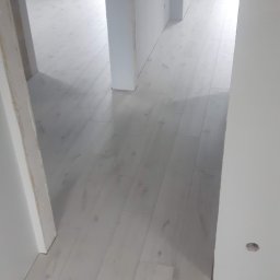 Krzysztof W. - Jasne panele podłogowe w nowym, minimalistycznym wnętrzu. Widok z perspektywy wejścia, z widocznymi fragmentami ścian i otworami drzwiowymi. Delikatne światło podkreśla strukturę paneli.