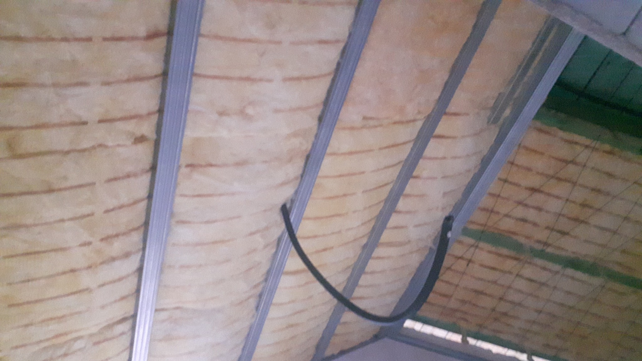 Izolacja poddasza wełną mineralną, widoczne metalowe profile i przewód. Ujęcie z dołu, ukazujące strukturę i detale izolacji termicznej.