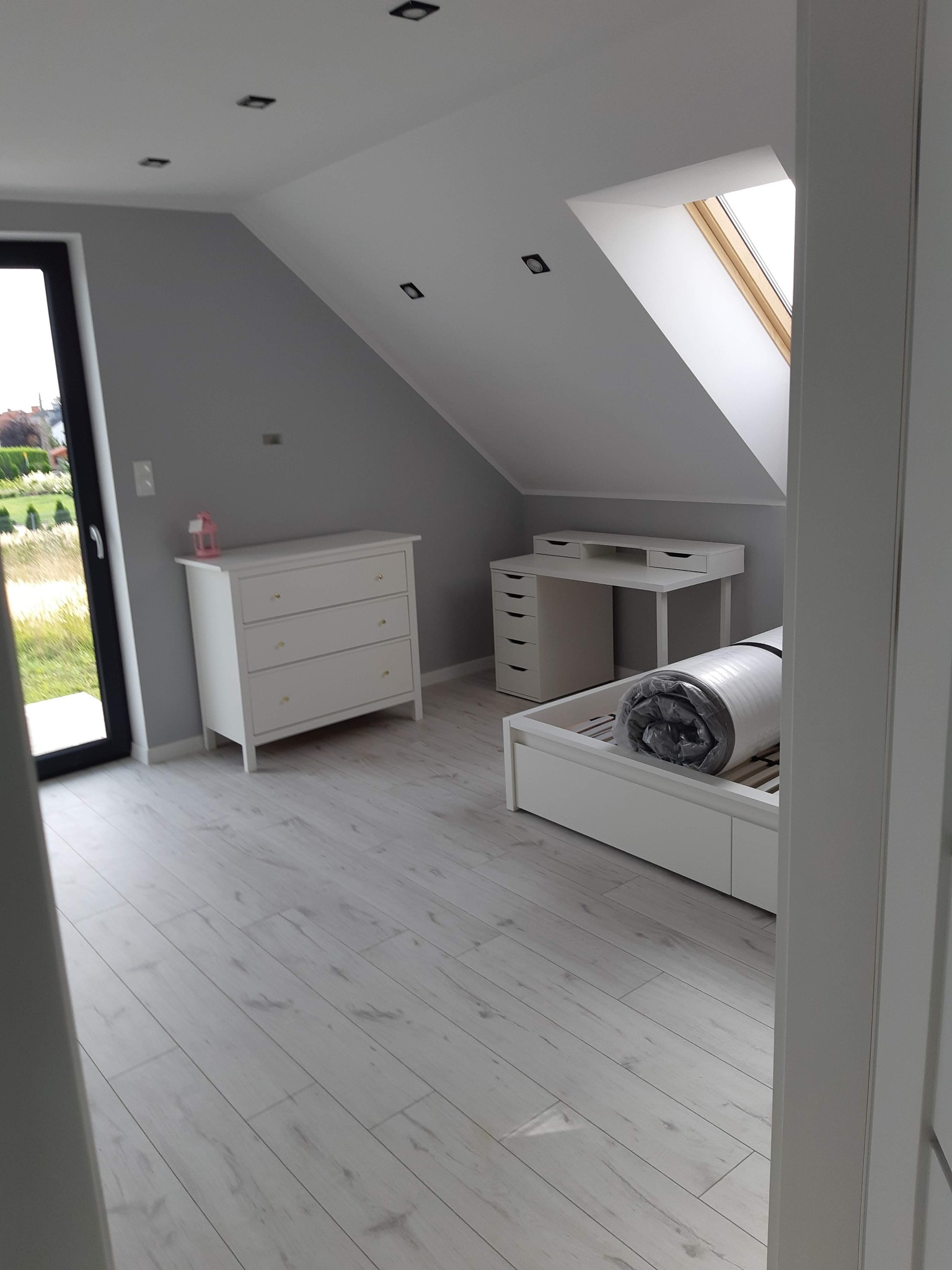 Jasne poddasze z oknem dachowym, białe meble (komoda, biurko, łóżko z materacem w folii) i panele podłogowe. Szare ściany, czarne lampy sufitowe.