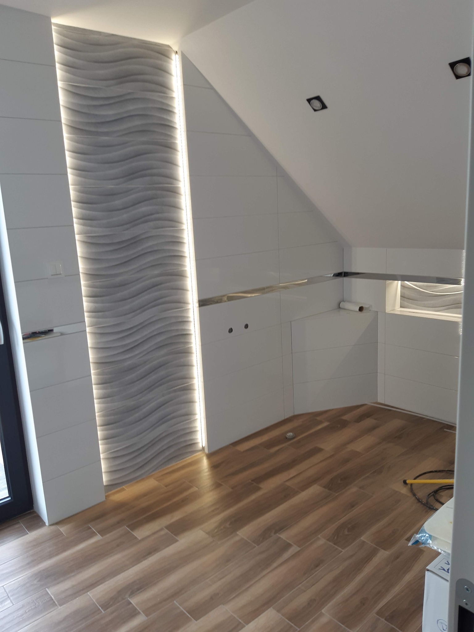 Wykończona łazienka na poddaszu z drewnopodobną podłogą, białą glazurą i dekoracyjnym panelem ściennym z podświetleniem LED. Widoczne detale wykończeniowe.