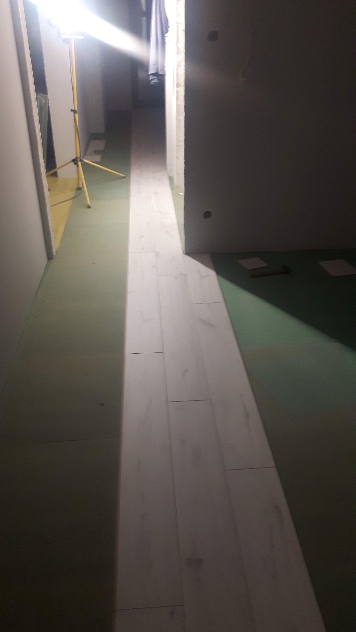 Ułożone panele podłogowe w korytarzu, widoczne narzędzia i lampa budowlana. Częściowo oświetlone, surowy stan ścian.