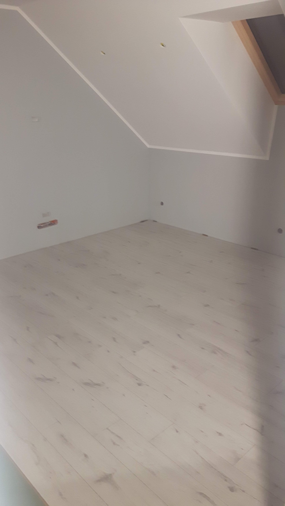 Jasne panele podłogowe w pokoju na poddaszu z oknem dachowym. Ściany pomalowane na szaro. Widoczne gniazdka elektryczne i otwory po instalacji.