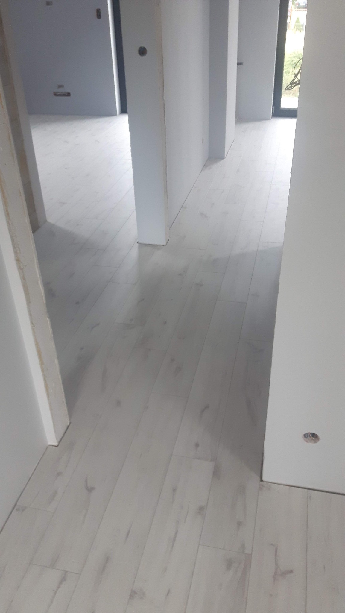 Jasne panele podłogowe w nowym, minimalistycznym wnętrzu. Widok z perspektywy wejścia, z widocznymi fragmentami ścian i otworami drzwiowymi. Delikatne światło podkreśla strukturę paneli.