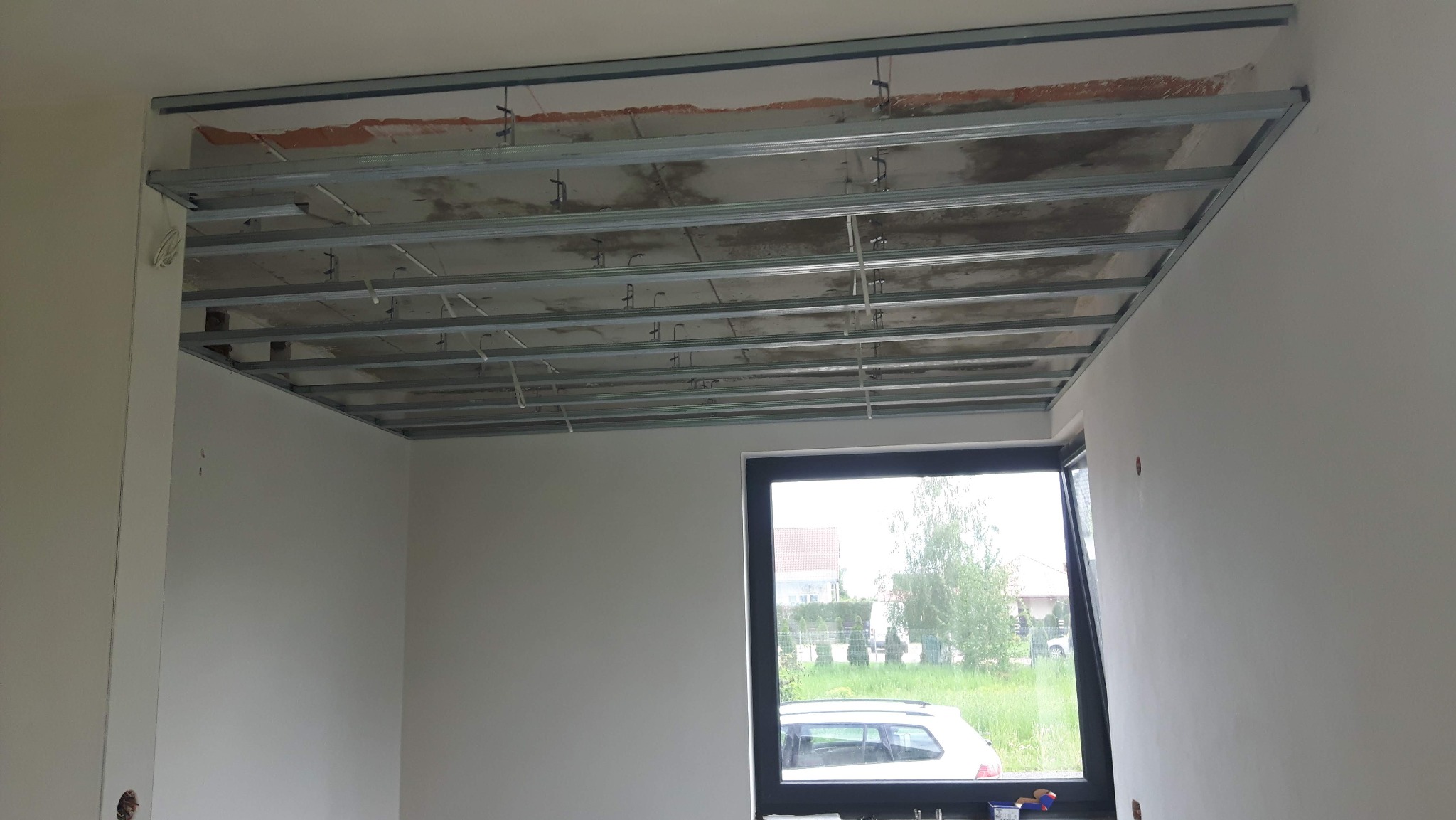 Stelaż sufitu podwieszanego z metalowych profili w surowym wnętrzu, widok z dołu na konstrukcję przed montażem płyt gipsowo-kartonowych, okno z widokiem na zieleń w tle.