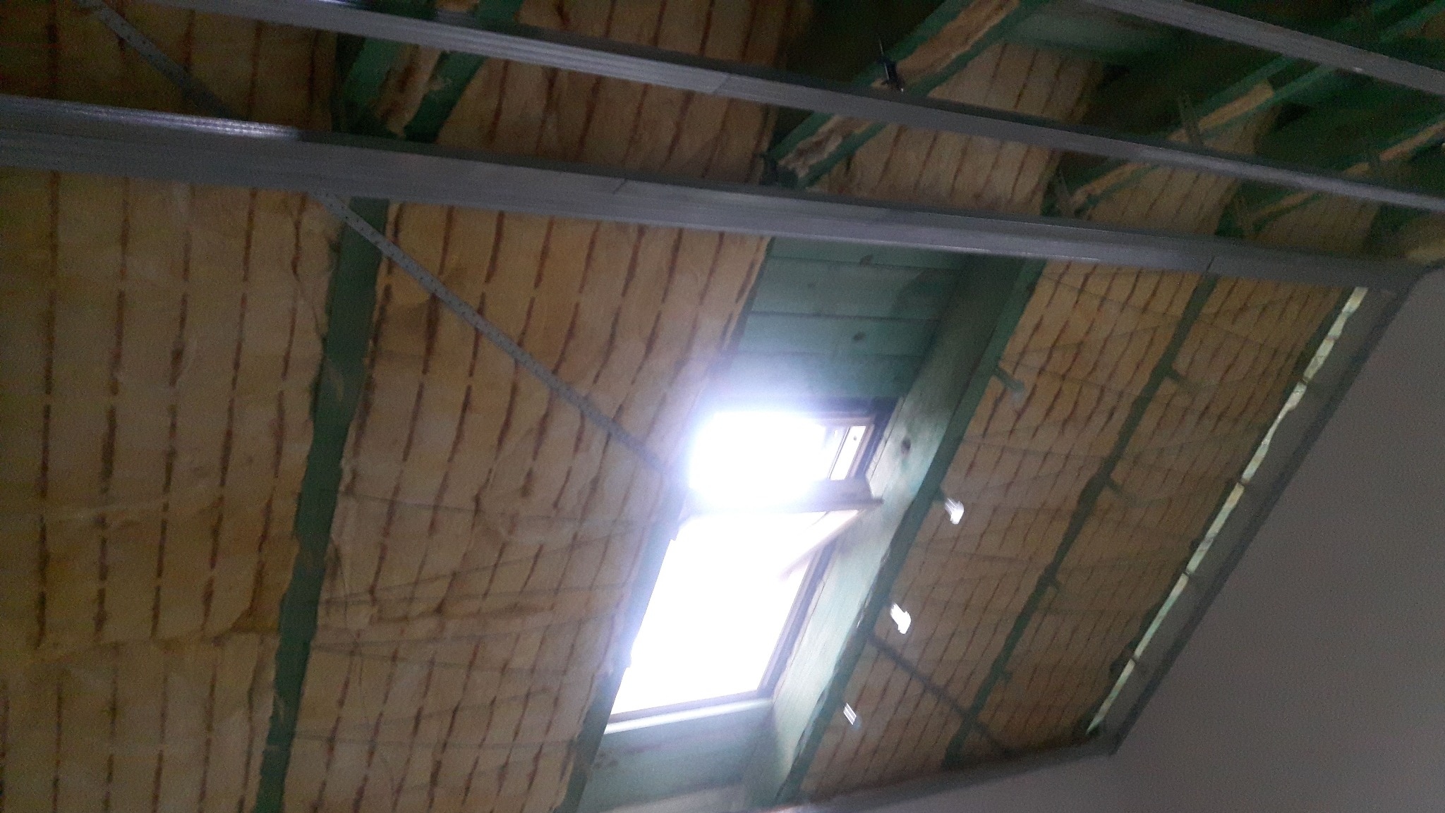 Ocieplenie poddasza wełną mineralną, widoczne elementy konstrukcji drewnianej i metalowe profile. Okno dachowe wpuszczające jasne światło.