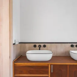 Nowoczesna łazienka z dwiema białymi umywalkami nablatowymi z czarnym obramowaniem, zamontowanymi na drewnianym blacie z szafkami, czarne baterie ścienne, beżowe płytki na ścianie i drewniane drzwi...