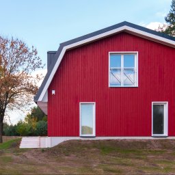 Anna Matus - Agent Nieruchomości - Czerwony budynek mieszkalny w stylu stodoły z białymi oknami i szarą dachówką, otoczony zielenią i drzewami w jesiennej scenerii.