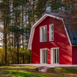Anna Matus - Agent Nieruchomości - Czerwony dom z białymi oknami i czarnym dachem, otoczony drzewami, z widocznym fragmentem tarasu i schodami z cegły.