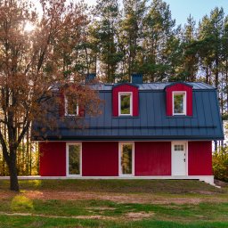 Anna Matus - Agent Nieruchomości - Nowoczesny, czerwony dom z ciemnoszarym dachem w otoczeniu drzew w Warszawie, widziany z niskiego kąta, z delikatnym światłem słonecznym przebijającym przez korony drzew.