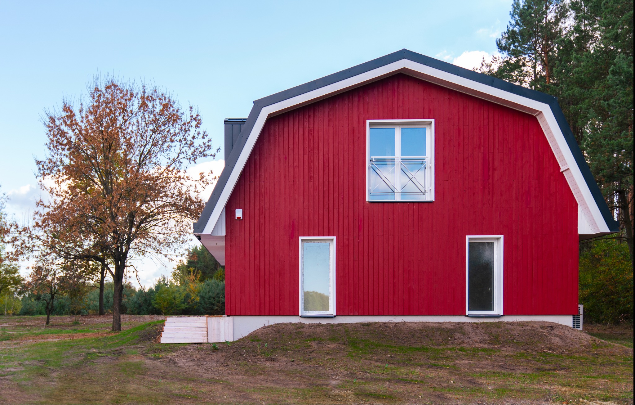 Czerwony budynek mieszkalny w stylu stodoły z białymi oknami i szarą dachówką, otoczony zielenią i drzewami w jesiennej scenerii.