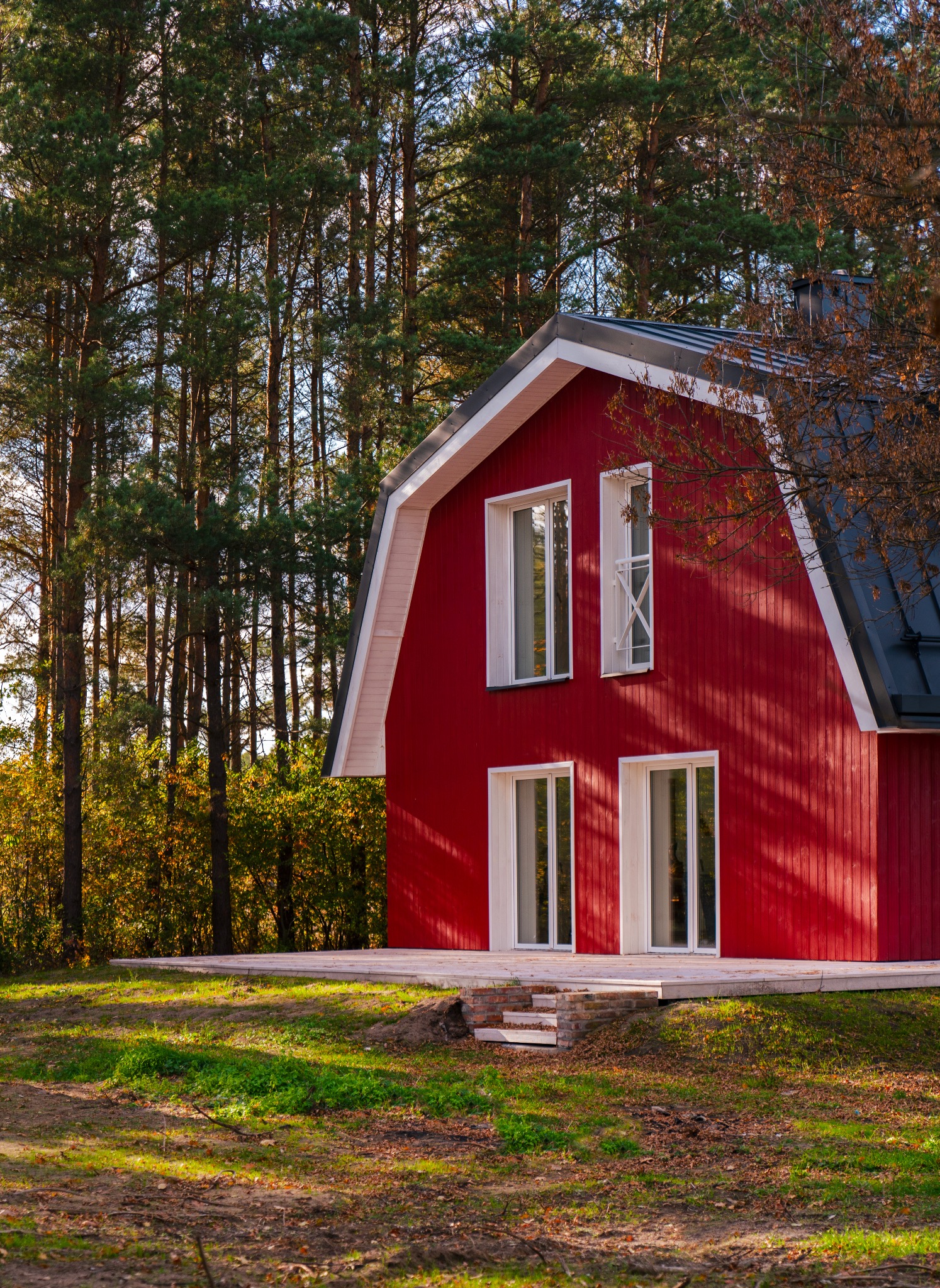 Czerwony dom z białymi oknami i czarnym dachem, otoczony drzewami, z widocznym fragmentem tarasu i schodami z cegły.