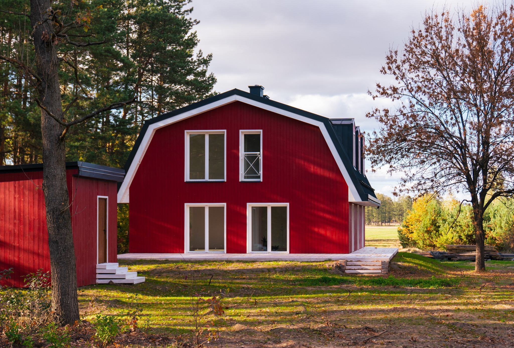 Czerwony dom w stylu stodoły z białymi oknami i czarnym dachem, otoczony drzewami w jesiennej scenerii.