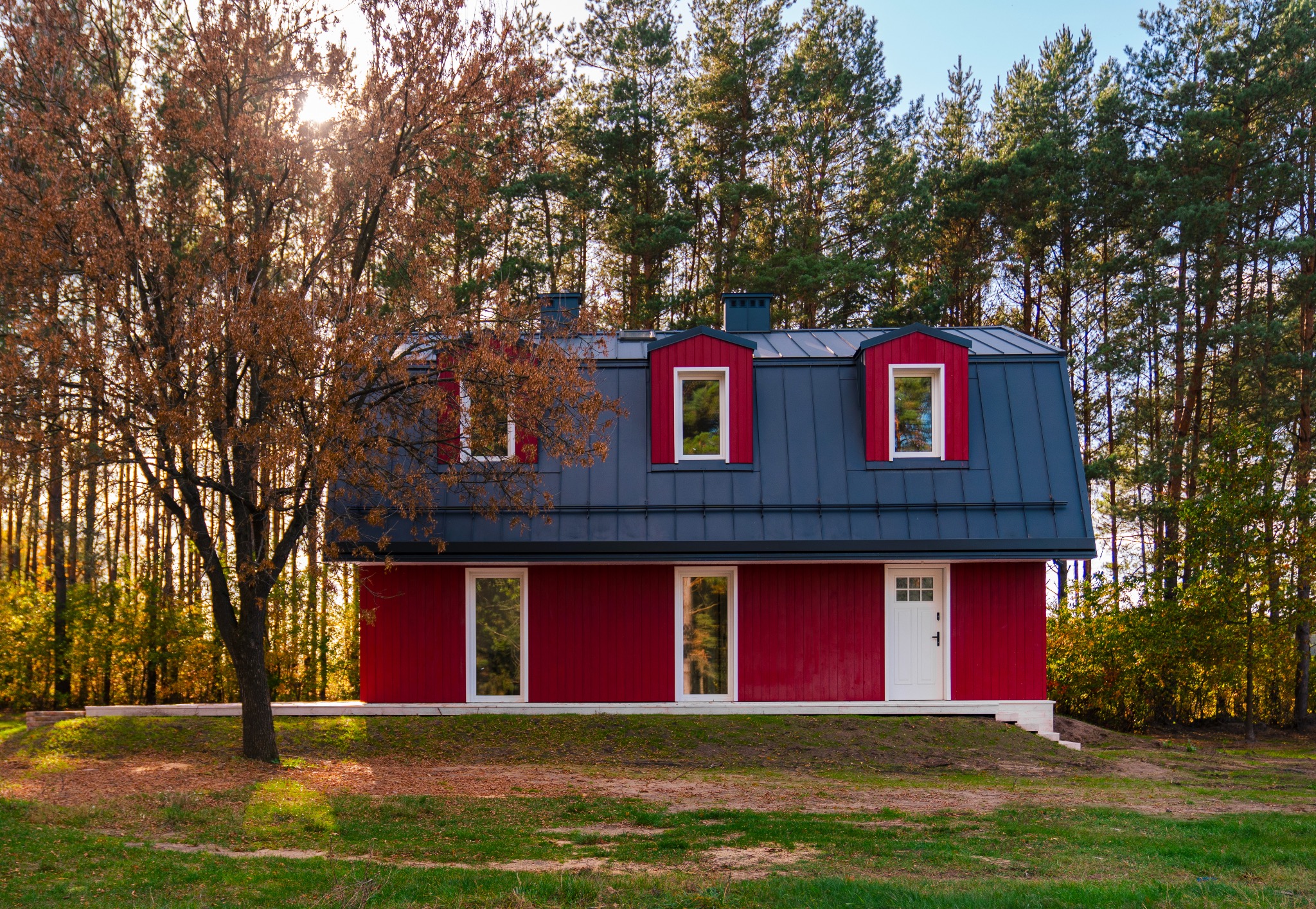 Nowoczesny, czerwony dom z ciemnoszarym dachem w otoczeniu drzew w Warszawie, widziany z niskiego kąta, z delikatnym światłem słonecznym przebijającym przez korony drzew.