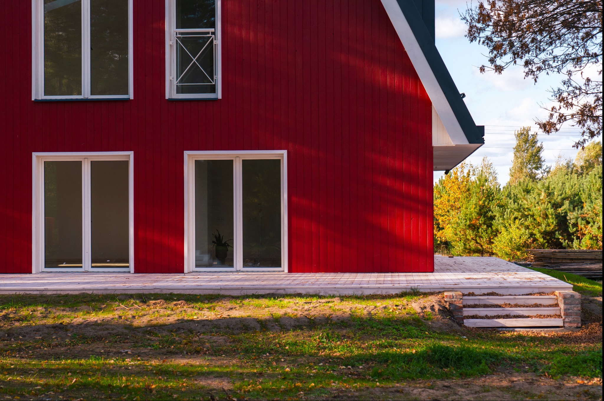 Czerwony, drewniany dom z białymi ramami okiennymi i schodami prowadzącymi do wejścia, otoczony jesienną zielenią.