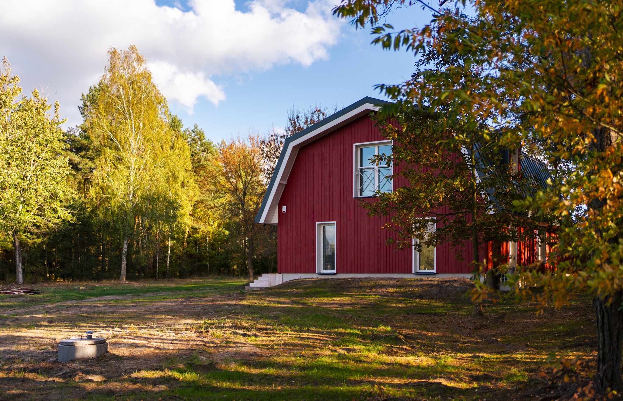 Czerwony, drewniany dom z dwuspadowym dachem, otoczony jesiennymi drzewami i trawnikiem; widoczny właz do studzienki kanalizacyjnej.