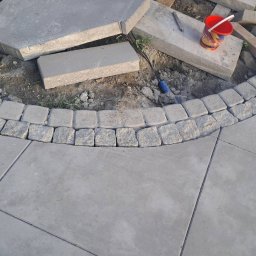 SolidCraft - Fragment placu z ułożoną częściowo kostką granitową tworzącą łuk, obok rozłożone płyty chodnikowe i czerwone wiaderko.