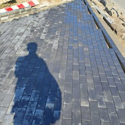 SolidCraft - Fragment nowej nawierzchni z ciemnoszarej kostki brukowej, z widocznym cieniem osoby fotografującej i fragmentem drogi w tle.