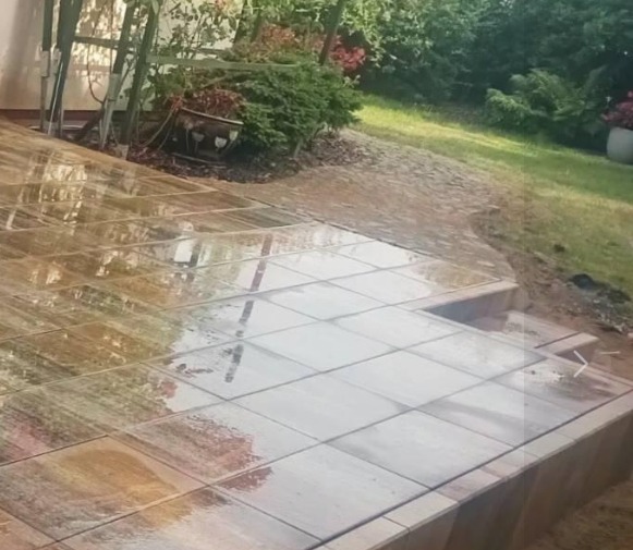 Świeżo ułożony taras z płytek imitujących kamień, z widocznymi schodkami i mokrą powierzchnią odbijającą otaczającą zieleń ogrodu.