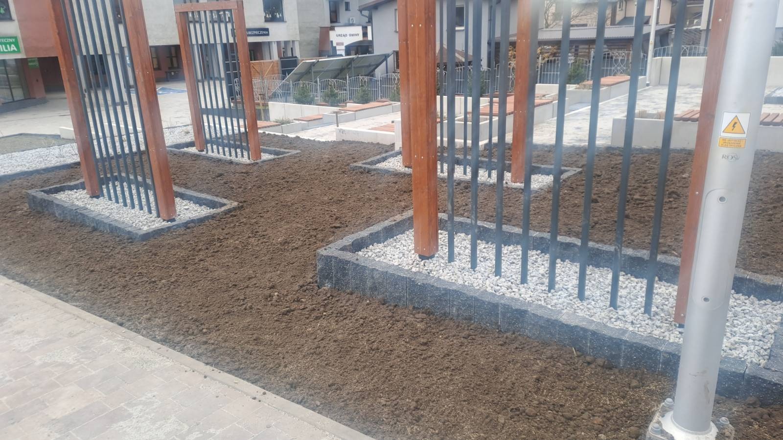 Przygotowany teren pod nasadzenia z obrzeżami z kostki brukowej i drewniano-metalowymi konstrukcjami, widoczny słup oświetleniowy z naklejką ostrzegawczą.