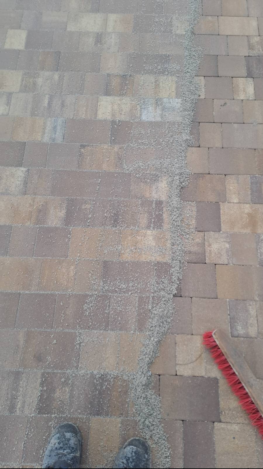 Układanie kostki brukowej: Wypełnianie szczelin piaskiem kwarcowym na brązowo-szarej kostce brukowej, widoczny fragment szczotki do zamiatania i buty robocze.