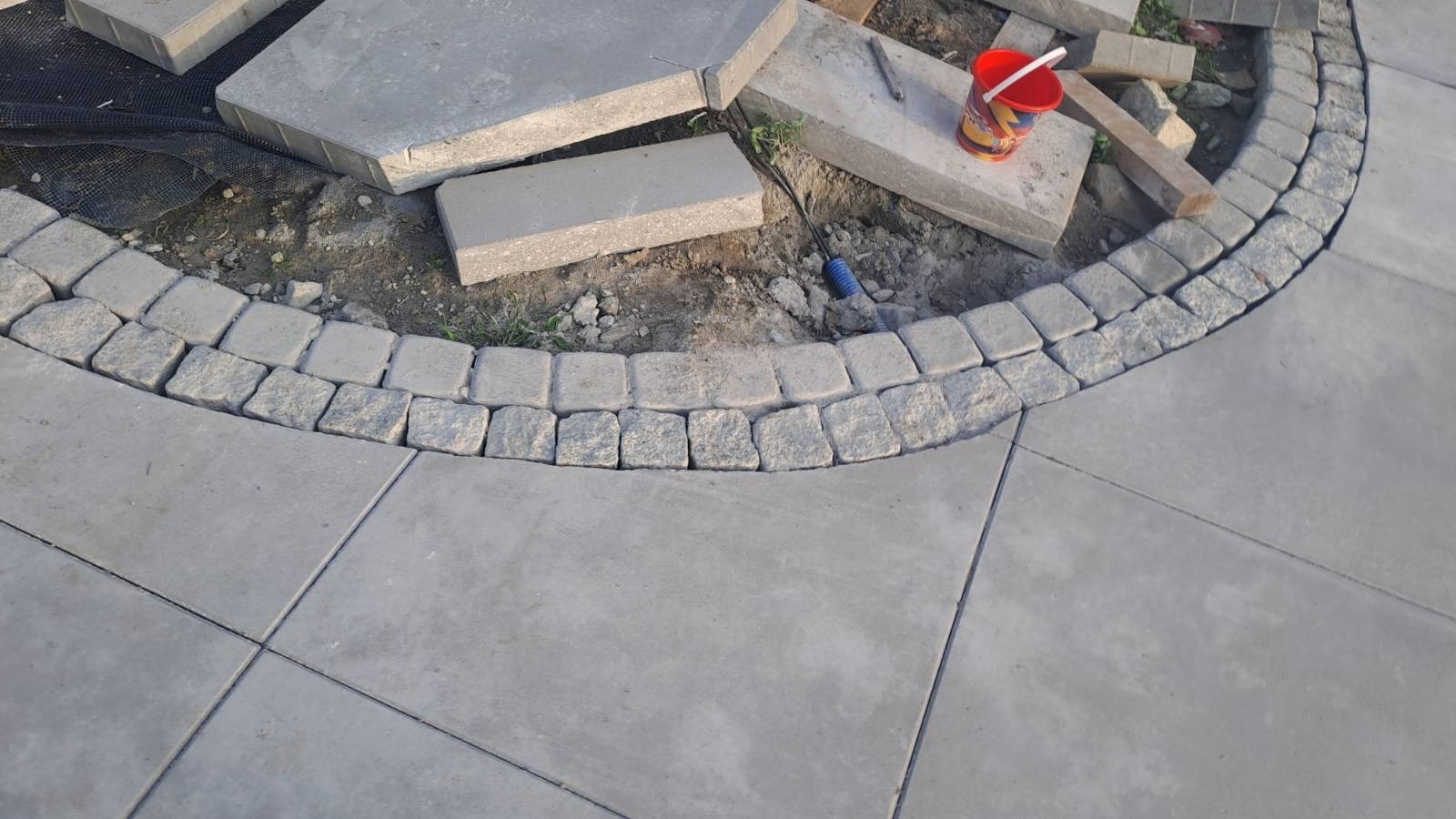 Fragment placu z ułożoną częściowo kostką granitową tworzącą łuk, obok rozłożone płyty chodnikowe i czerwone wiaderko.