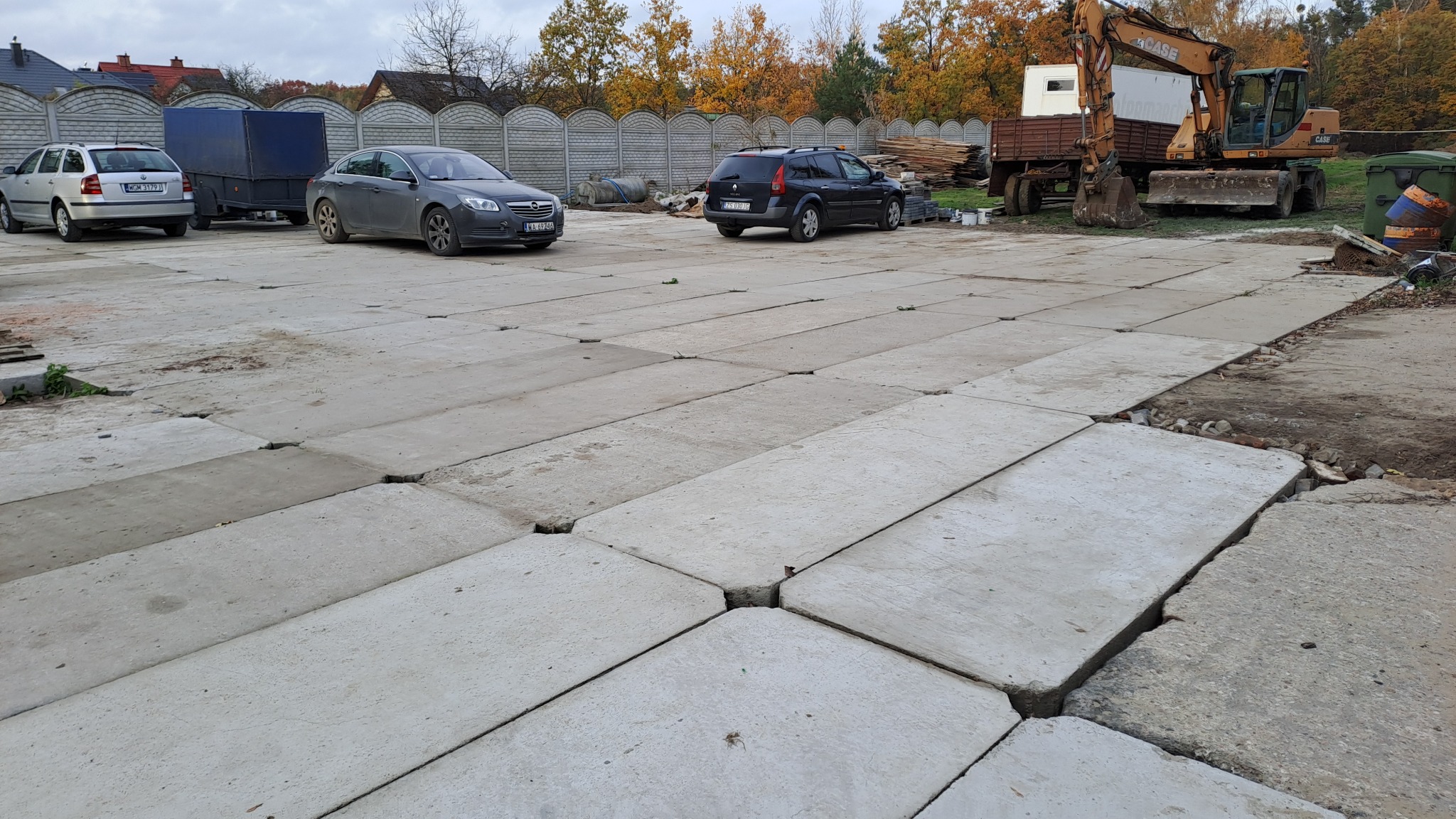 Ułożona nawierzchnia z betonowych płyt na terenie budowy, z widocznymi samochodami, przyczepą i koparką w tle, częściowo zniszczona na krawędziach.