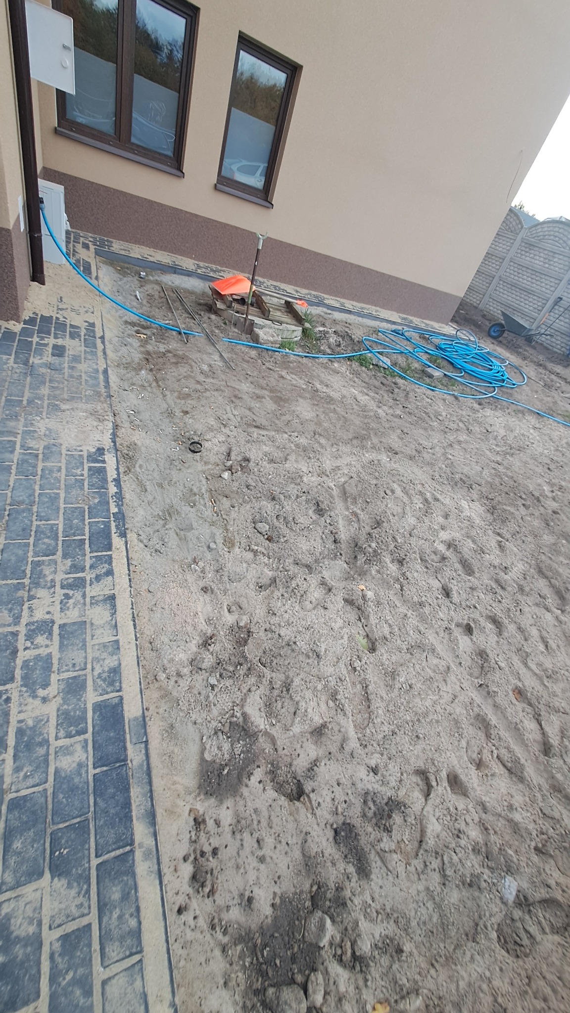 Przygotowanie podłoża pod układanie kostki: wyrównany teren z widocznymi śladami stóp, narzędzia pracy (szpadel, poziomica), niebieski wąż ogrodowy, fragment ułożonej kostki brukowej przy ścianie...