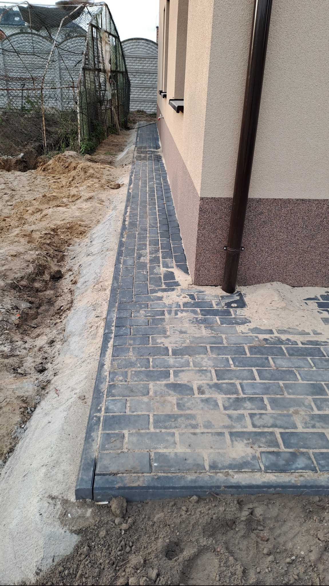 Ułożona ścieżka z szarej kostki brukowej, obrzeża z ciemnoszarego betonu, piasek na powierzchni, rynna przy ścianie budynku, widok z boku.