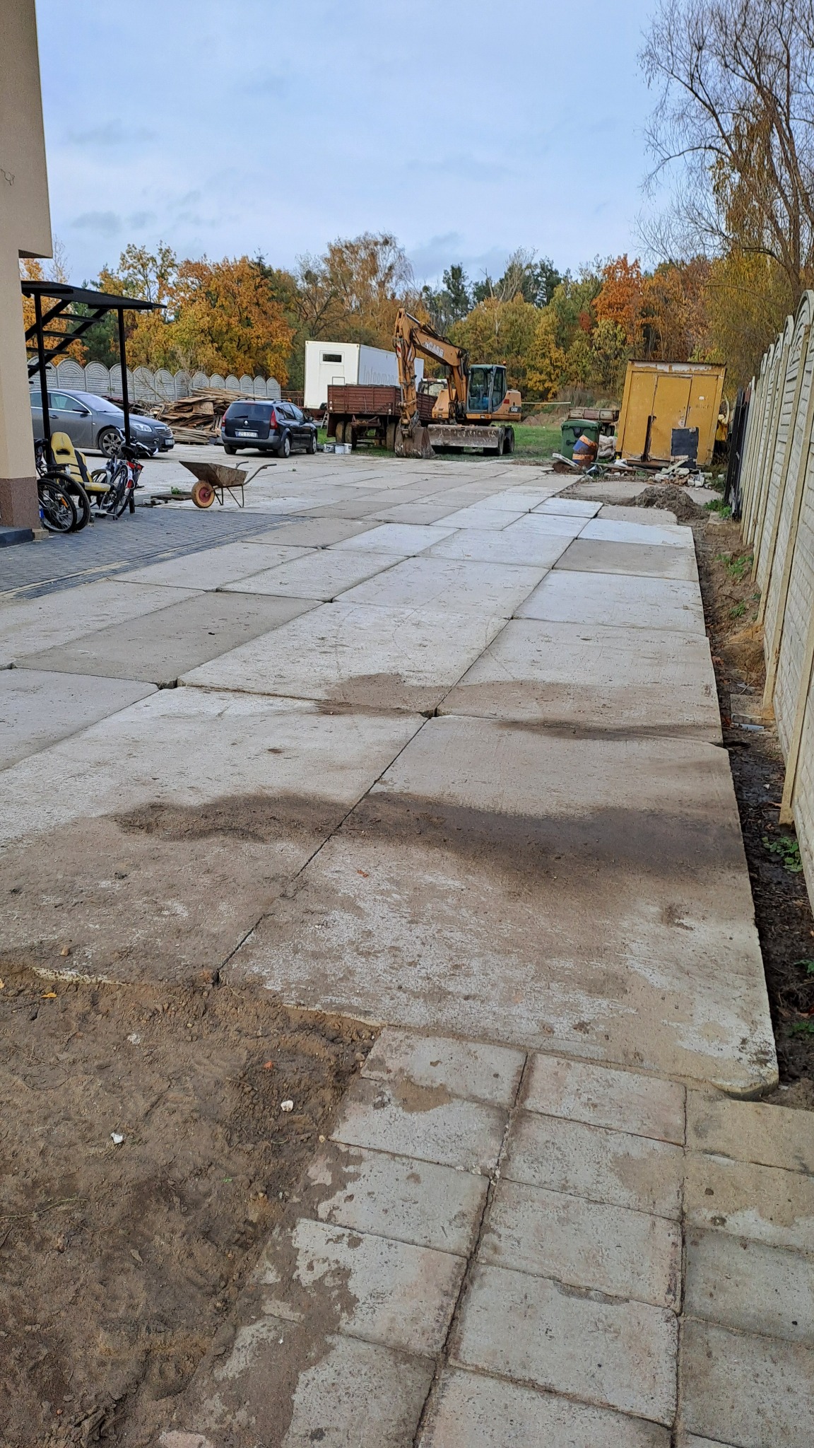 Rozległy plac wyłożony betonowymi płytami w trakcie prac, z widocznym sprzętem budowlanym i ziemią przy krawędziach, na tle jesiennego krajobrazu z drzewami o barwnych liściach.