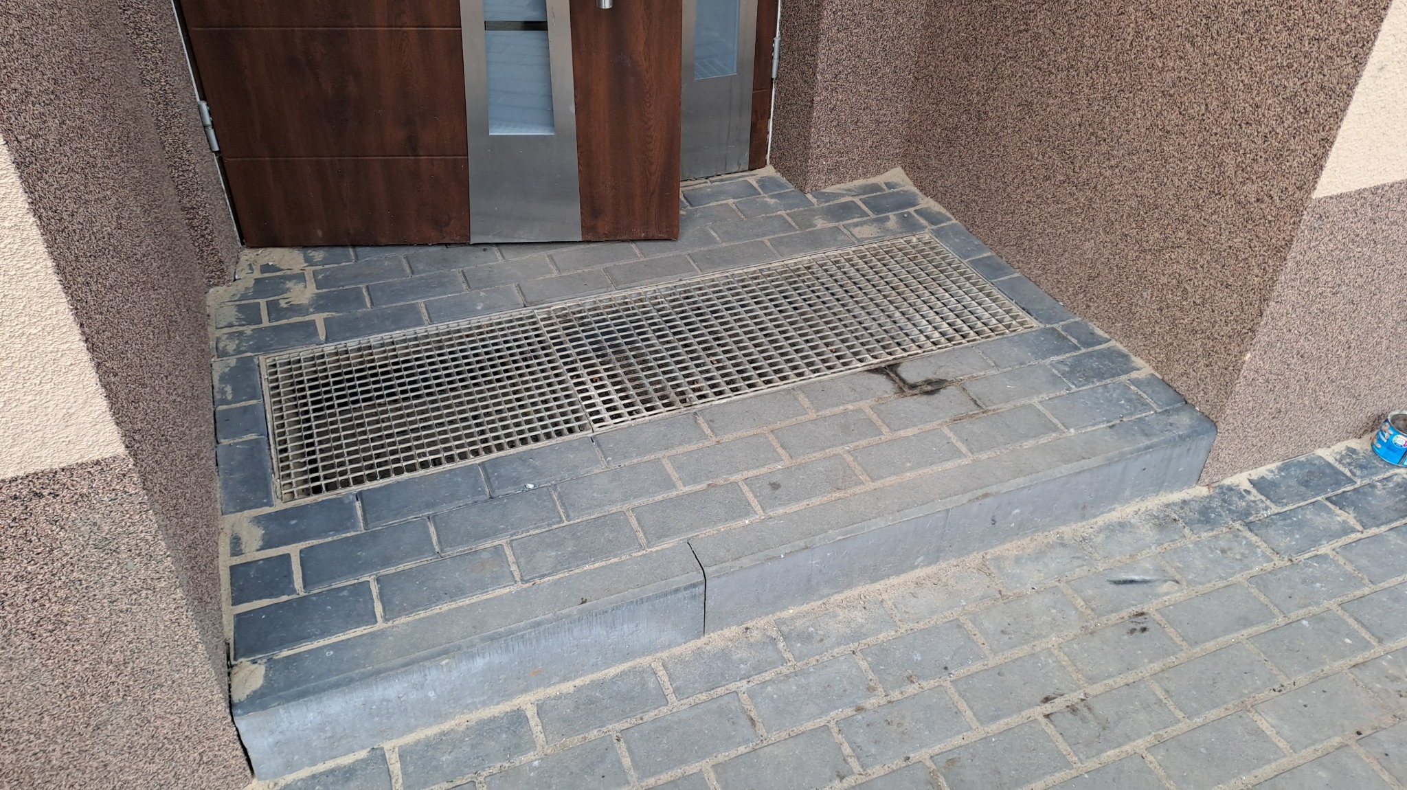 Ułożona szara kostka brukowa przed wejściem do budynku z kratką odpływową, widoczne zabrudzenia piaskiem i resztki zaprawy.