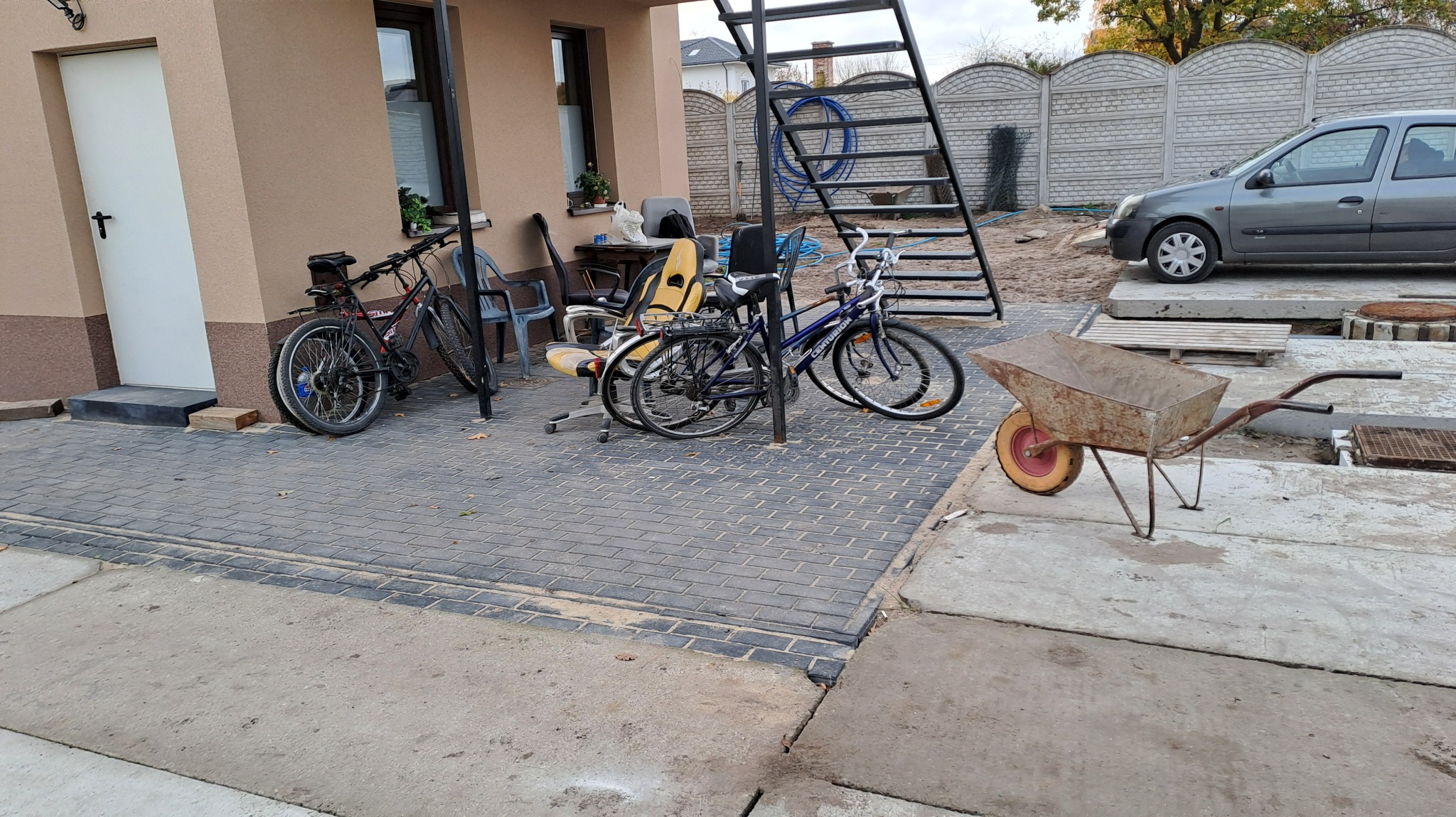 Ułożona szara kostka brukowa przed domem z zaparkowanymi rowerami, taczką i samochodem w tle.