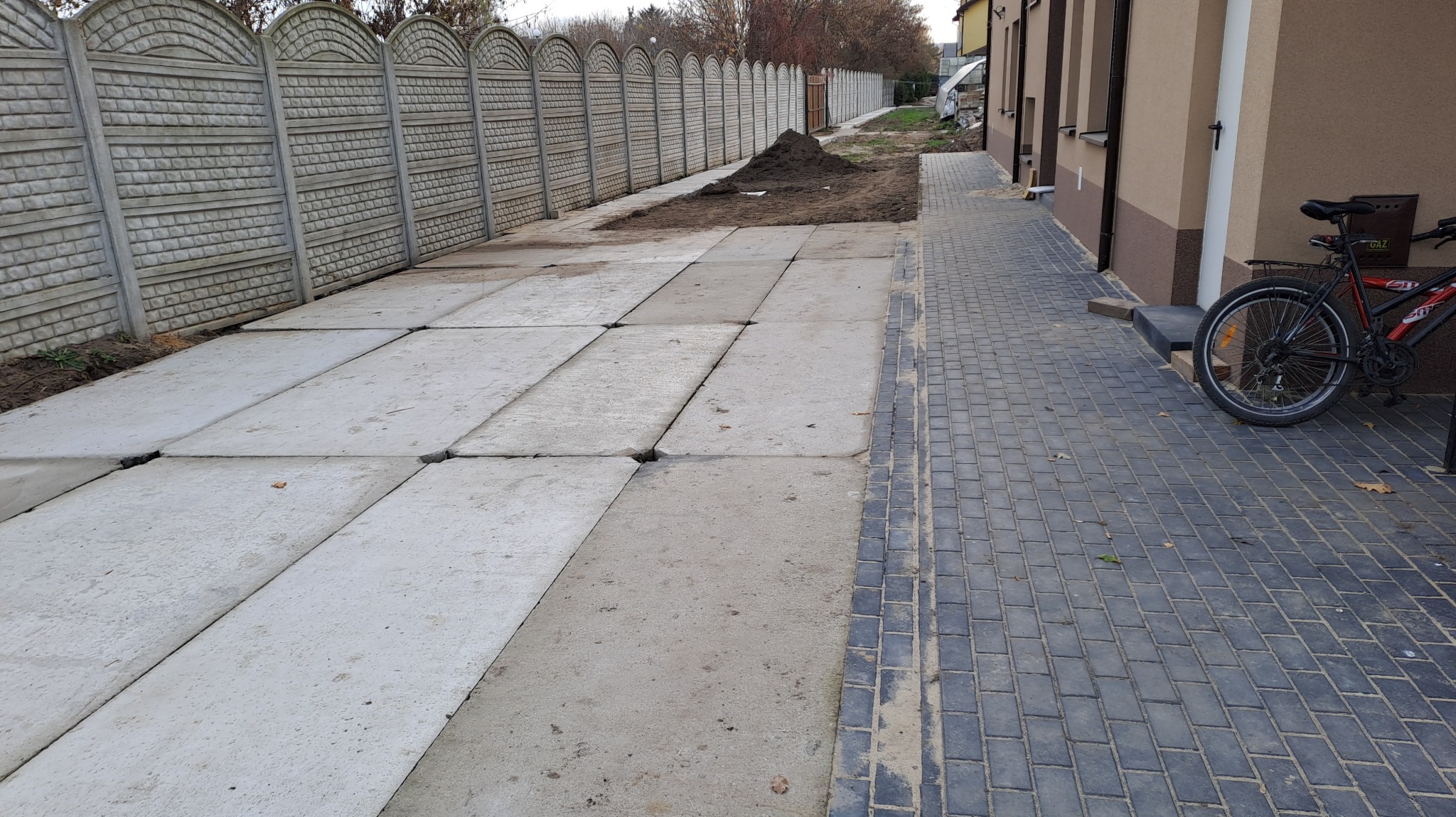 Układanie nawierzchni z betonowych płyt i kostki brukowej, widoczny fragment ogrodzenia z prefabrykatów betonowych oraz rower oparty o ścianę budynku.