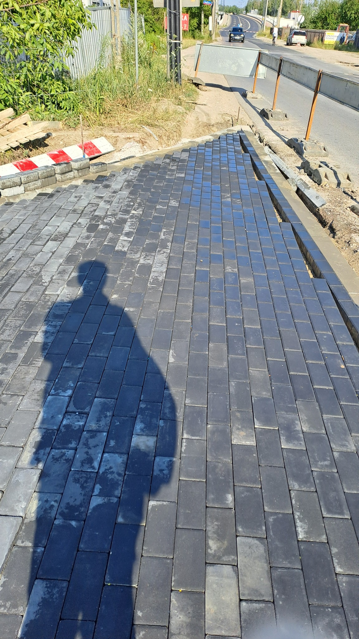 Fragment nowej nawierzchni z ciemnoszarej kostki brukowej, z widocznym cieniem osoby fotografującej i fragmentem drogi w tle.