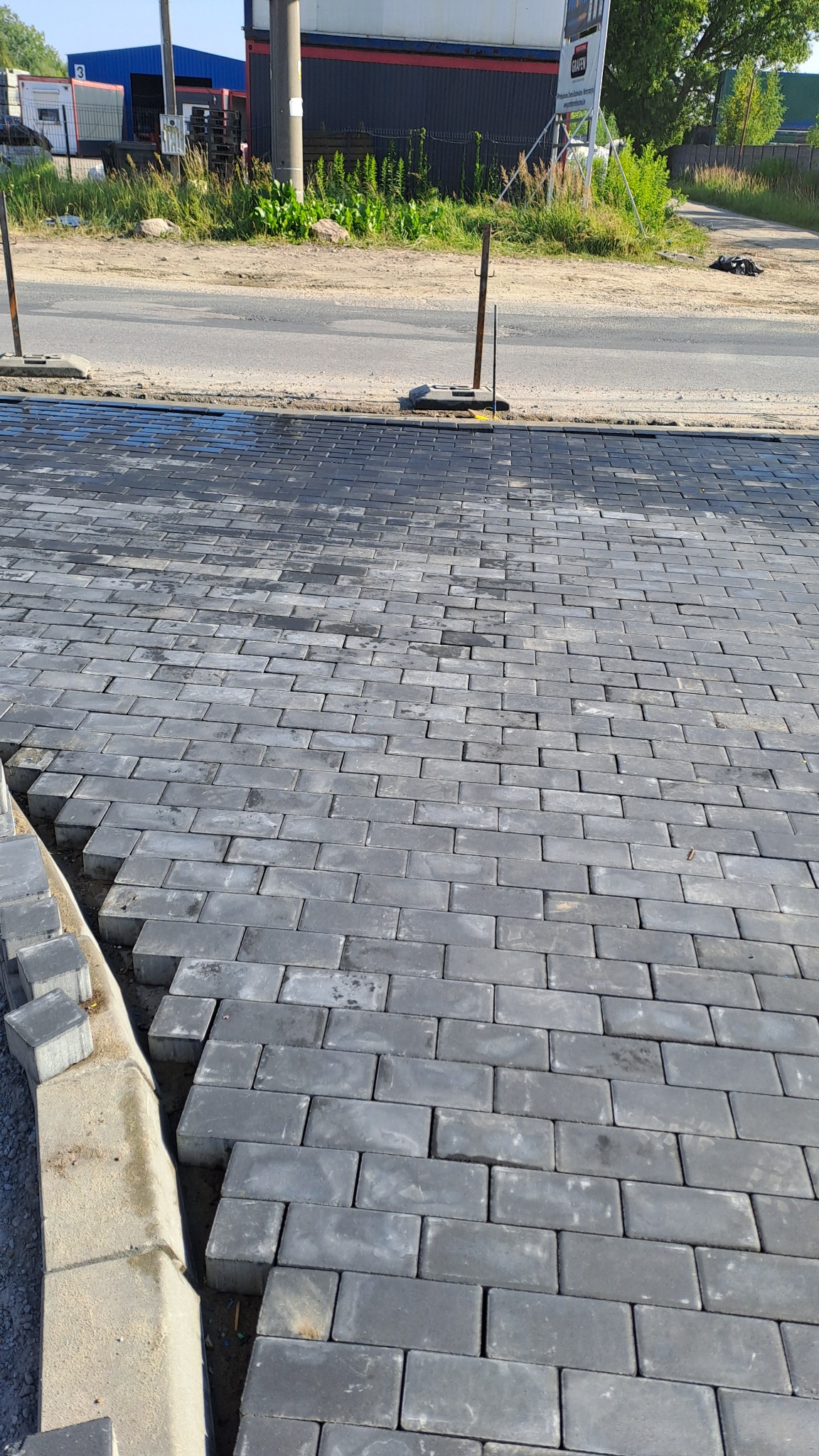 Układanie szarej, prostokątnej kostki brukowej na podjeździe, widoczne niezakończone prace przy krawężniku.