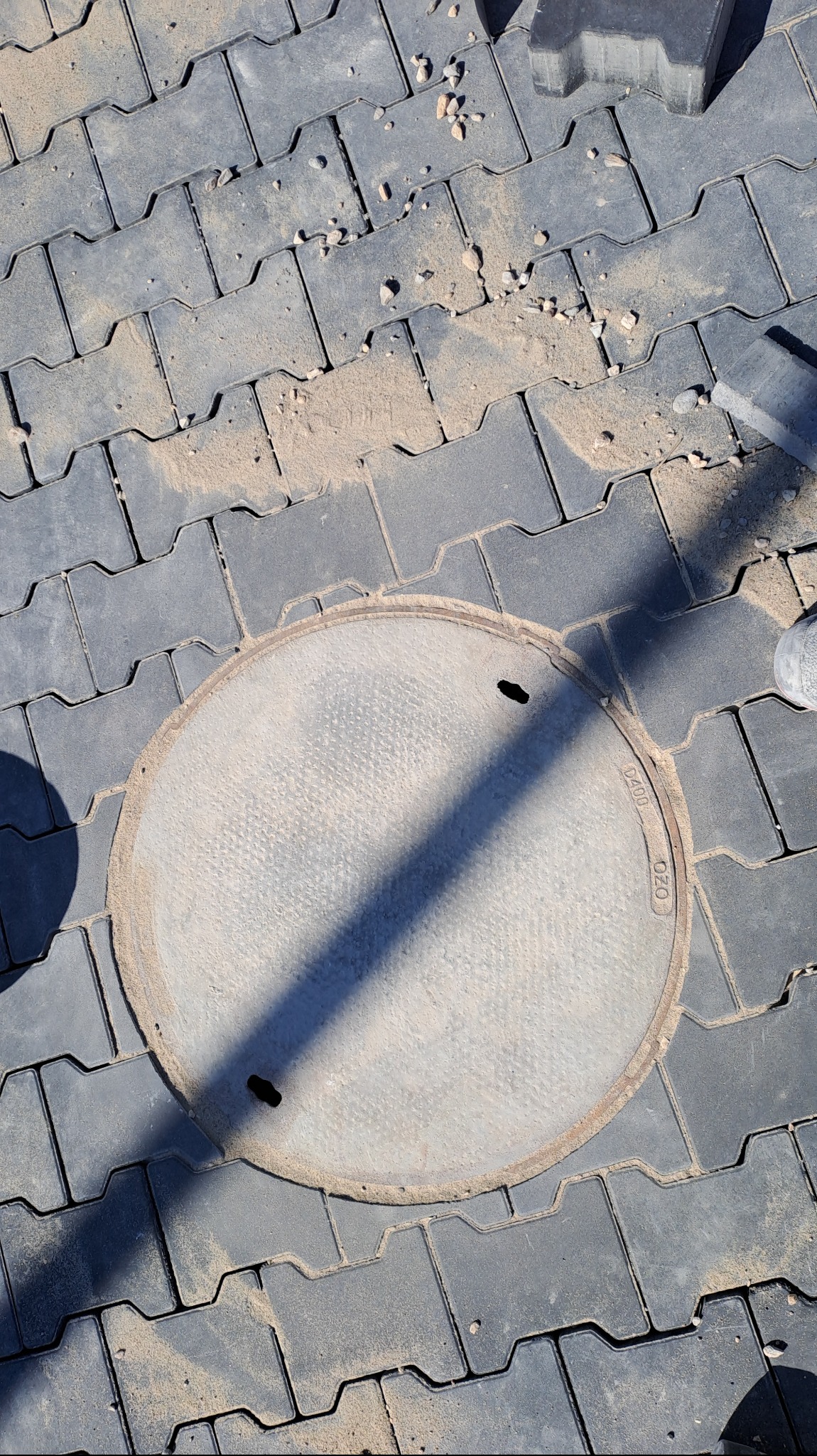 Szare, ułożone kostki brukowe wokół okrągłej studzienki kanalizacyjnej z widocznymi śladami piasku i drobnych kamieni na powierzchni.